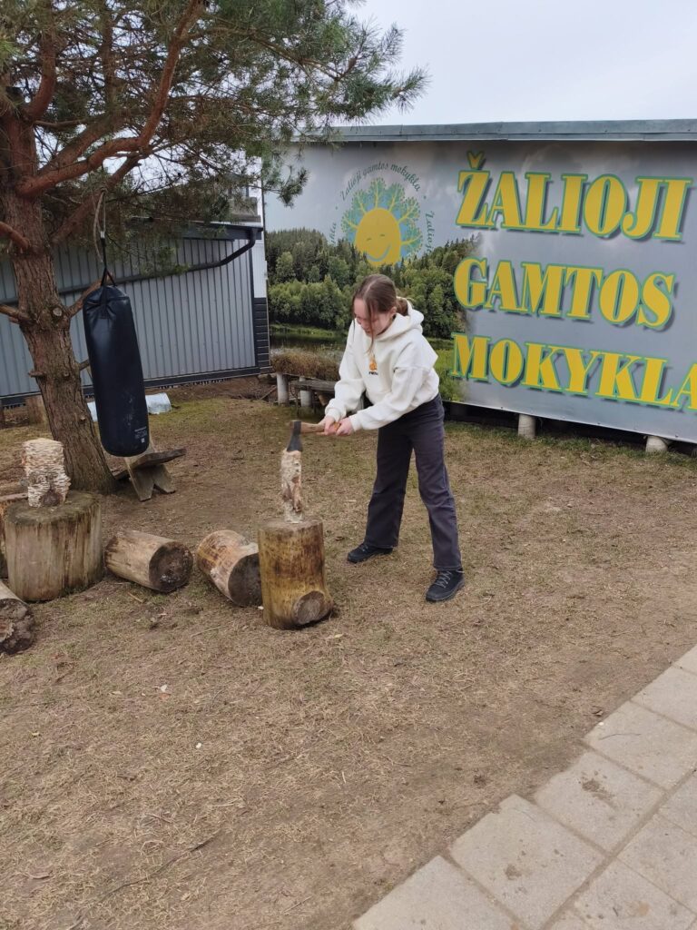Žaliosios gamtos mokyklos vaikas lauke skaido malką ir mokosi savarankiškumo bei atsakomybės
